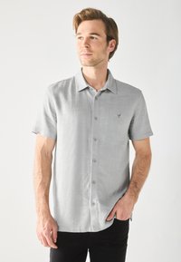 Camisa de manga corta de color gris claro con una tela texturizada, cierre de botones y un pequeño logotipo bordado en el pecho. Corte casual.