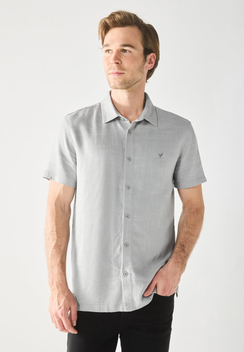 Camisa de manga corta de color gris claro con una tela texturizada, cierre de botones y un pequeño logotipo bordado en el pecho. Corte casual.