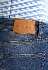 Bakvy av blå denimjeans med en brun läderetikett som står "PIER ONE" och en blå och vit randig skjorta instoppad.