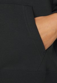 Main insérée dans la poche avant d'un sweat à capuche en coton noir, montrant la texture du tissu et les détails de la couture.