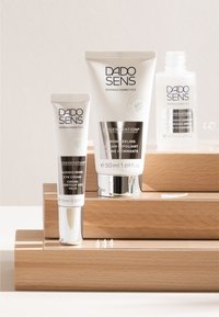 Three DADO SENS skincare products: a white tube, a white jar with silver accents, and a transparent bottle, displayed on wooden risers.