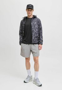Sort og grå mønstret letvægtsjakke, sort T-shirt, grå shorts og grå træningssko med hvide sokker, stående mod en ensfarvet baggrund.
