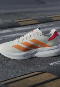 Baskets blanches avec des rayures orange et un détail rouge au talon reposant sur l'asphalte près d'un ralentisseur jaune et noir.