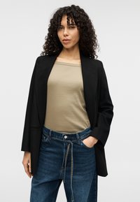 Μαύρο σακάκι με δομημένους ώμους, συνδυασμένο με μπεζ αμάνικη μπλούζα και μπλε, φαρδιά τζιν. Τα κατσαρά μαλλιά προσθέτουν σε μια casual εμφάνιση.