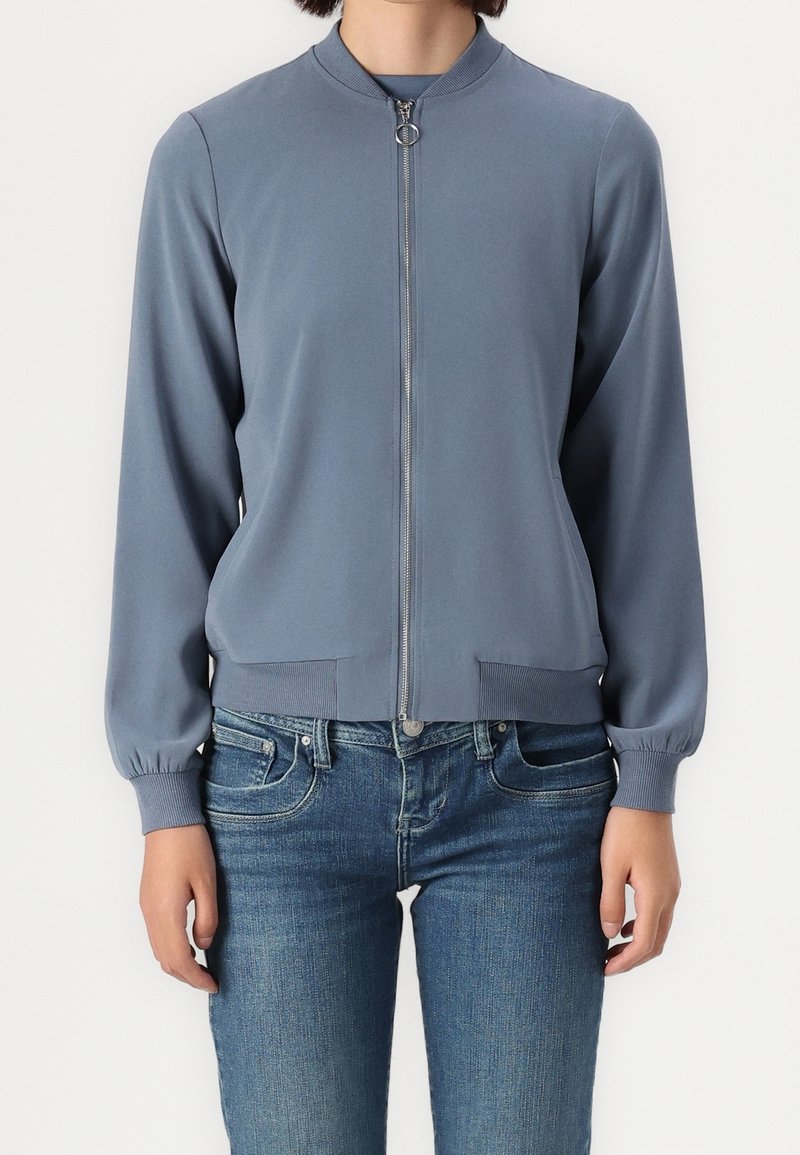 Ljusblå zip-up bomberjacka gjord av mjukt tyg, med rund krage, ribbade manschetter och två framfickor.
