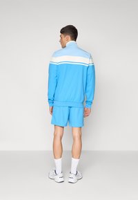 Casaco de desporto azul claro com riscas brancas e azul escuro, combinado com calções a condizer. Apresenta um colarinho alto e punhos com ribetes.