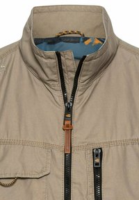 Beige Jacke mit Stehkragen, ausgestattet mit einem durchgehenden Reißverschluss, zwei Fronttaschen und blauer gemusterter Innenauskleidung. Strukturierter Stoff mit kontrastierenden Akzenten.