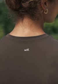 Sötétbarna, testre szabott pulóver kerek nyakkivágással, finom textúrával és a felső háton egy fehér nyomott "self." logóval.