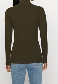 Langärmliger, enganliegender Rollkragenpullover aus dunkelolivgrünem geripptem Stoff, mit hohem Kragen und strukturierten vertikalen Streifen. Kombiniert mit hellblauen Jeans.