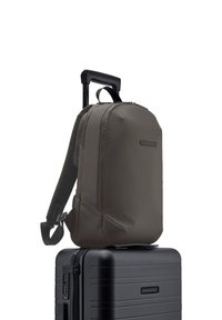 Sac à dos vert olive avec des bretelles rembourrées et un compartiment principal zippé, reposant sur une valise à roulettes noire avec une finition texturée.