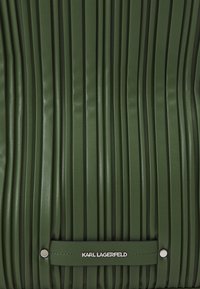 Bolso de cuero verde con pliegues verticales, que presenta una etiqueta de logo plateada y detalles a presión. Textura suave con una forma estructurada.