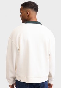 Light beige sweatshirt with a green collar, featuring a relaxed fit and ribbed cuffs. Includes a small logo tag at the hem.
