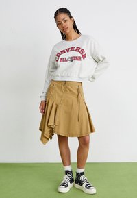 Sudadera gris con el texto "CONVERSE ALL STAR" en rojo, falda asimétrica caqui con cremalleras y bolsillos, y zapatillas negras de caña alta.