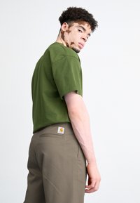 Ung mand med vitiligo iført olivengrønt t-shirt og brune Carhartt bukser, som ser over skulderen mod en hvid baggrund.