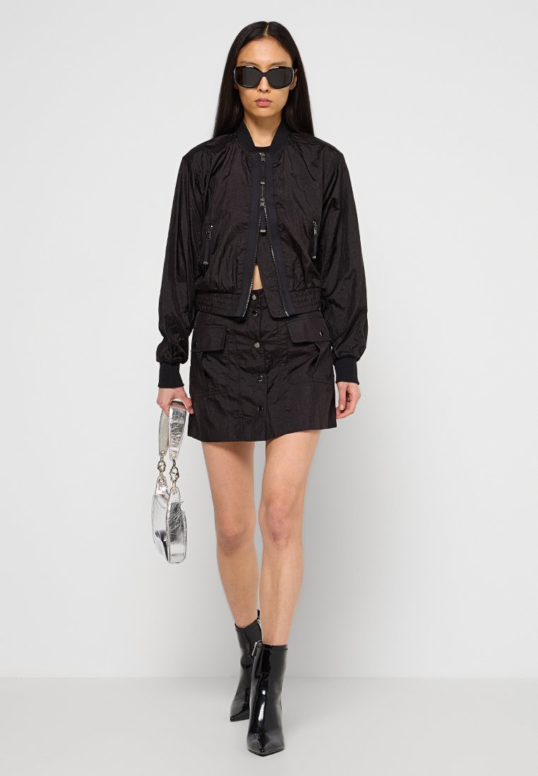 Schwarze Bomberjacke mit Reißverschluss und elastischen Bündchen, kombiniert mit einem schwarzen Rock mit Knöpfen vorne. Accessoires umfassen eine transparente Handtasche und Sonnenbrille.
