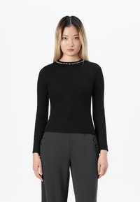 Young woman with straight blonde hair wearing a fitted black long-sleeve top with pearl detail and dark wide-leg pants standing against white background.