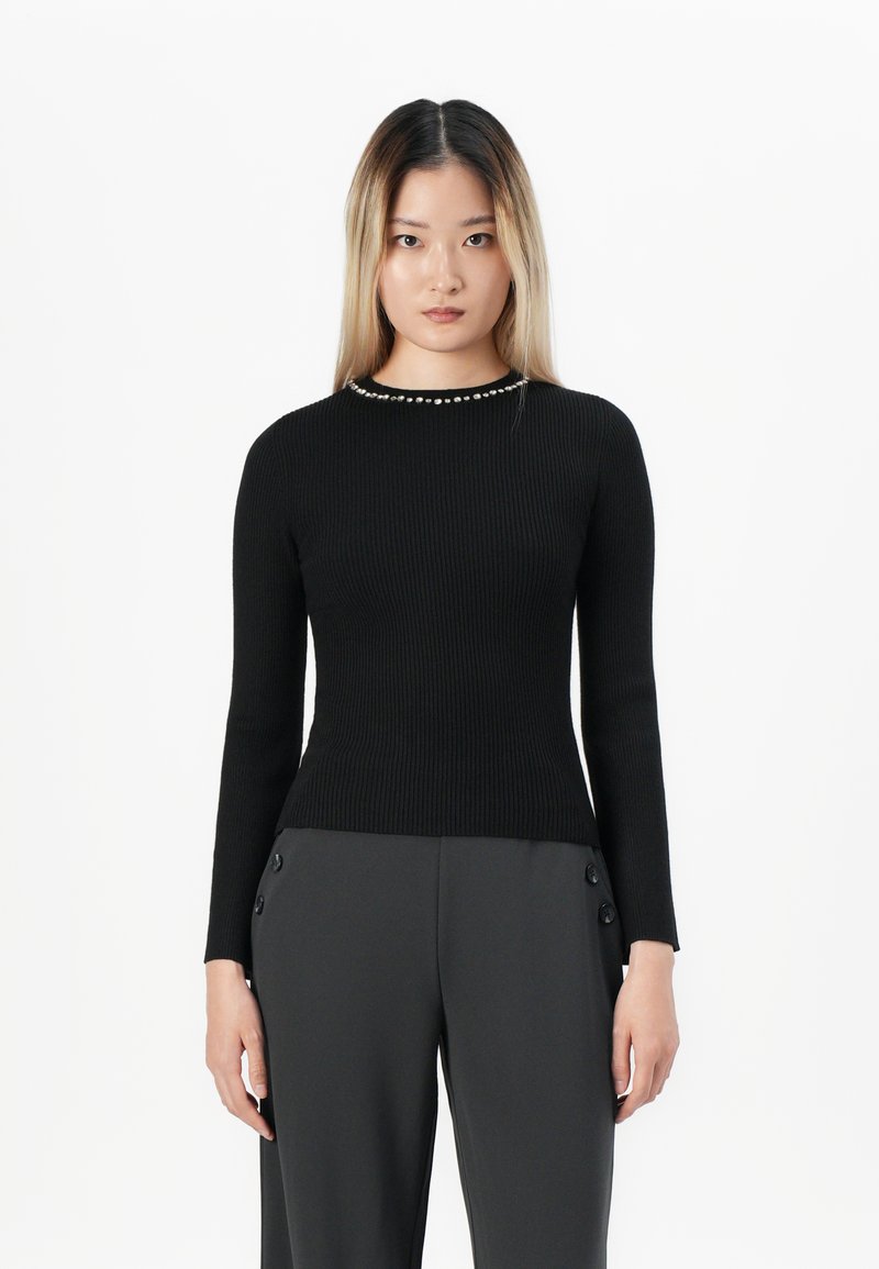 Young woman with straight blonde hair wearing a fitted black long-sleeve top with pearl detail and dark wide-leg pants standing against white background.