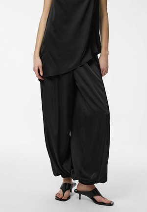 Person som har på seg løse svarte satengbukser med elastiske mansjetter og svarte åpne kitten heel-mules mot en hvit bakgrunn.