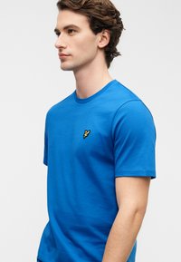 Μπλε βαμβακερό μπλουζάκι με στρογγυλή λαιμόκοψη, με μικρό κίτρινο κεντημένο λογότυπο στο αριστερό στήθος και κοντά μανίκια.