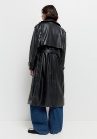Personne portant un trench-coat en cuir noir ceinturé à la taille, associé à un jean large bleu et des chaussures noir et blanc, se tenant de dos.