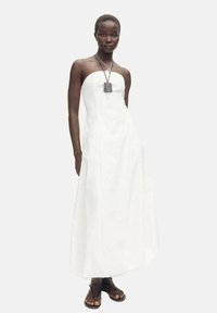 Robe maxi blanche sans bretelles avec un corsage ajusté, une jupe évasée et des poches latérales. Le mannequin est accessorisé avec un collier superposé et des sandales.