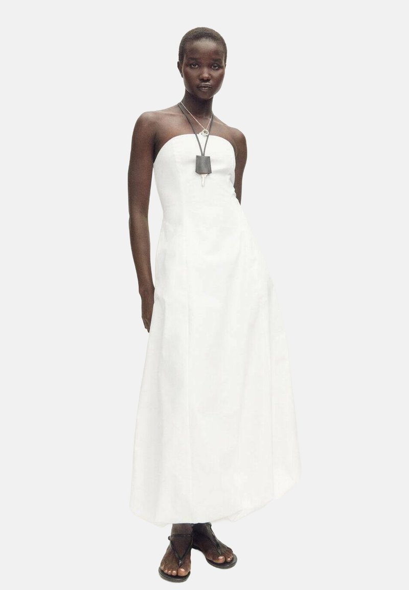 Robe maxi blanche sans bretelles avec un corsage ajusté, une jupe évasée et des poches latérales. Le mannequin est accessorisé avec un collier superposé et des sandales.