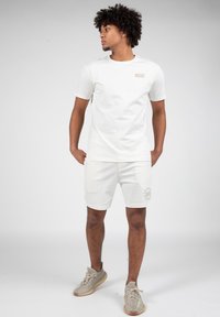 Hvid bomulds t-shirt og matchende shorts med en diskret logo på brystet og et grafisk motiv på benet, parret med sneakers i neutrale farver.