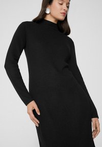Schwarzes geripptes Strickkleid mit langen Ärmeln und hohem Kragen, das eine figurbetonte Silhouette und eine glatte Textur aufweist. Silberne runde Ohrringe.