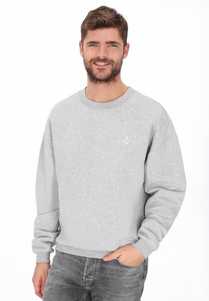 Glimlachende man met kort bruin haar die een lichtgrijze trui draagt met een klein wit ankerlogo, poserend met één hand in zijn zak.