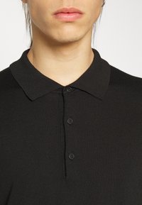 Černé polo tričko vyrobené z žebrovaného materiálu, se plochým límečkem, zapínáním na tři knoflíky a hladkým povrchem. Bez viditelných vzorů nebo akcentů.