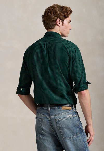 Man with curly hair wearing a dark green checkered long-sleeve shirt and blue jeans, standing with his back facing the camera.