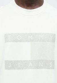 Λευκή φούτερ μπλούζα με ανάγλυφο γραφικό εκτύπωμα σε ανοιχτό γκρι και το κείμενο «TOMMY JEANS» στο κέντρο στο κάτω μέρος. Στρογγυλή λαιμόκοψη.