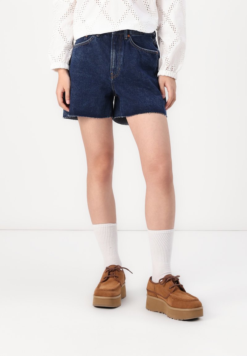 Jeansshorts, mörkblå, med en något fransad nederkant, två framfickor. Bärs med vita knästrumpor och bruna platåskor.