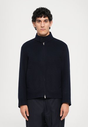 Marineblå zip-up jakke med høj krave, der har to sidelommer og et glat, tekstureret stof. Minimalistisk design uden synlige mønstre.