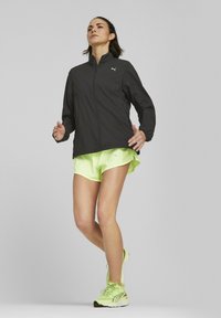 Sort atletisk jakke med høj krave, parret med neongule shorts. Jakken har en frontlynlås og subtil branding.