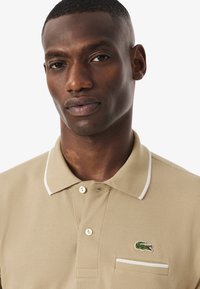 Beige polo-shirt med en texturerad tyg, vit krage, och en broderad grön krokodil-logotyp på bröstfickan.