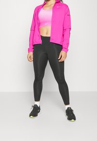 Chaqueta ligera rosa sobre un sujetador deportivo rosa, combinada con leggings negros y zapatillas deportivas negras. Materiales suaves con detalles de logotipo.