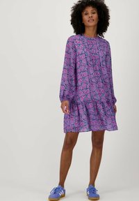 Paarse bloemenjurk met lange mouwen en een gerimpelde zoom. De stof heeft een patroon van roze en zwarte bloemen. Gekleed met blauwe sneakers.