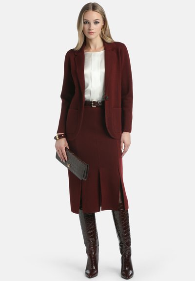 Woman wearing a burgundy blazer, matching skirt with front slits, white blouse, holding a brown clutch, and tall crocodile-pattern boots.