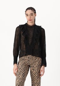 Zwarte doorzichtige blouse met ruches langs de halslijn en voorkant, stippenpatroon en kanten accenten. Gecombineerd met zebra print broeken.