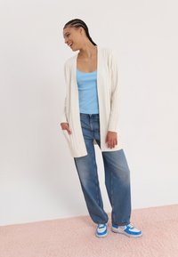 White knit cardigan, blue tank top, and loose denim jeans. Sneakers feature blue and white accents. Soft pink background.