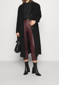 Cappotto lungo nero con un design sartoriale, abbinato a pantaloni di pelle bordeaux e stivali neri alla caviglia. L'outfit è completato con una borsa nera.