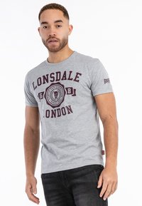 Pilka medvilninė marškinėliai su vyno spalvos grafiniu spaudiniu ir užrašu „Lonsdale London“. Trumpomis rankovėmis, apvaliu iškirpimu, prigludusio silueto ir logotipu ant rankovės.