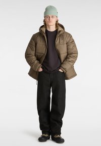 Pufferjack in olijfgroen, met een fleece gevoerde capuchon, zwarte shirt eronder, baggy zwarte jeans en zwarte schoenen met rubberen zolen.