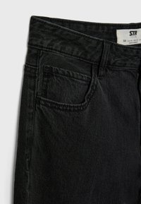 Gros plan d'un jean en denim noir montrant la ceinture, la poche avant, la boucle de ceinture et l'étiquette de taille à l'intérieur de la ceinture.