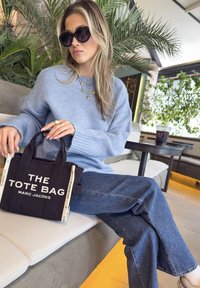 Černá bavlněná taška s bílým textem "TAŠKA MARC JACOBS", krátkými uchy a kontrastními popruhy. Modelka v světle modrém svetru a džínách.