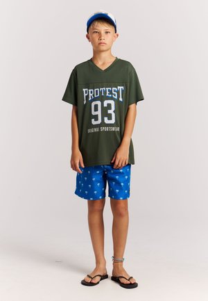 Groen mesh T-shirt met V-hals en 'PROTEST 93' opdruk, gecombineerd met blauwe korte broek met witte palmboomprint, zwarte slippers.