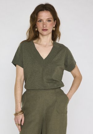 Woman with wavy auburn hair wears gold hoop earrings, olive green V-neck short sleeve top, matching high-waisted pants, and gold bracelets.