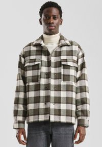 Geruite jas in groen en wit, gemaakt van dikke stof. Voorzien van twee borstzakken, knoopsluiting en een kraag.