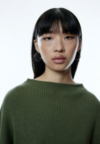 Grüner, gerippter Strickpullover mit weiter Ausschnitt. Das Model hat glattes schwarzes Haar und trägt goldene Creolen. Einheitlicher weißer Hintergrund.